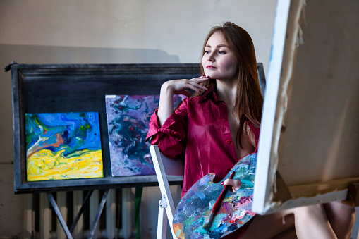 Lovely woman painter in red shirt painting with oil paints on canvas in art lab, thought looking away. Portrait of pretty woman artist sits in studio. Artisanal lifestyle concept. Copy ad text space