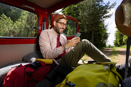 Happy male backpacker text messaging on cell phone while relaxing in his van.
