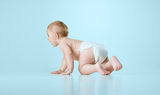 Curious and cheerful baby, toddler in diaper crawling on floor against light blue studio background. Concept of childhood, newborn lifestyle, health, happiness, care. Copy space for ad