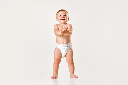 Portrait of little baby girl, toddler in diaper standing and clapping hands against white studio background. Happy child. Concept of childhood, newborn lifestyle, happiness, care. Copy space for ad