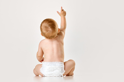 Back view image of little baby, child, toddler sitting in diaper against white studio background. Curious baby. Concept of childhood, newborn lifestyle, happiness, care. Copy space for ad