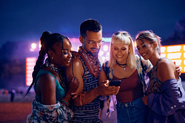 Young happy friends using mobile phone while attending live performance during summer music festival.