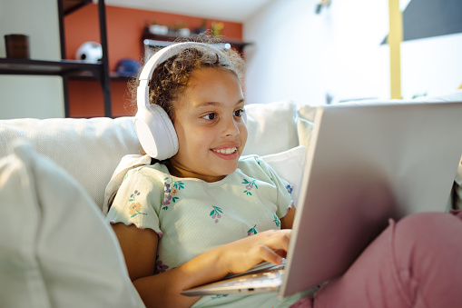 A young Latin American girl wearing headphones is sitting on a sofa and typing on her laptop with a smile.