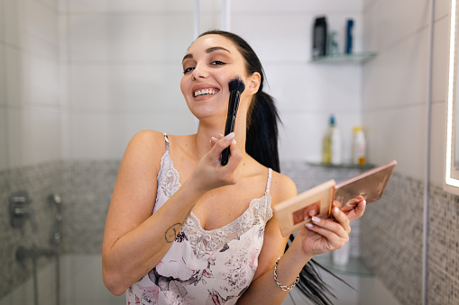 Embracing a seamless makeup application process, this young woman effortlessly enhances her features using a brush, transforming her look with grace and ease in the privacy of her bathroom