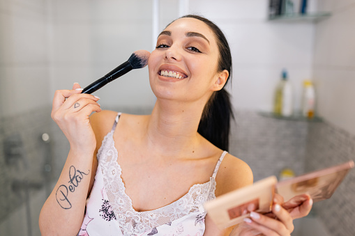 Showcasing her precision and finesse, this young woman flawlessly applies makeup with a brush, meticulously perfecting her look as she gazes into the bathroom mirror