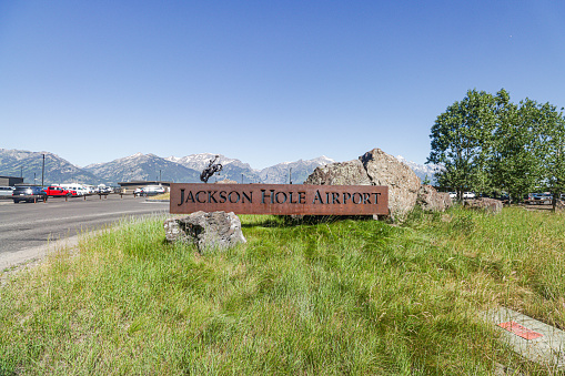 Cars with number plates visible behind the welcome sign for Jackson Hole Airport in Teton County, Wyoming. BEHIND the place sign is a 4,000 pound sculpture of a cowboy in a rodeo known as 'Battle of Wills', as designed by Bart Walter in 2016 to welcome visitors to the Cowboy State. The sign and the statue are two separate structures. This airport, a national monument, is the only US airport to be located in a national park - Grand Teton National Park in this case.
