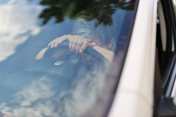 疲れた年配の女性が車のハンドルに寄りかかって、フロントガラス越しに見えた。 - owner women car rear view mirror ストックフォトと画像