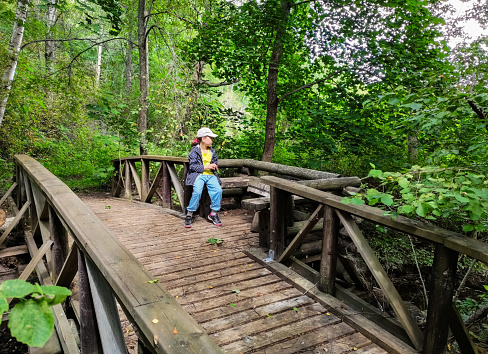An 8-year-old girl sits on an old abandoned wooden bridge, with a recreation area, across a narrow river in the forest.