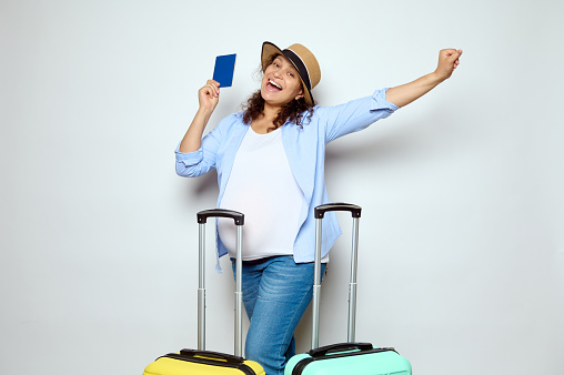 Happy pregnant woman in straw hat, white t-shirt and casual jeans, holding passport, expressing positive emotions, traveling abroad, going on vacation, isolated on white. Air travel. Tourism. Holidays