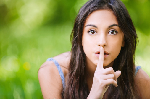 Young beautiful dark-haired woman has put forefinger to lips as sign of silence, against green summer garden.