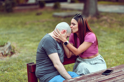 Balding Male Due To Chemotherapy Getting Head Kisses By Wife While Relaxing Together In Park