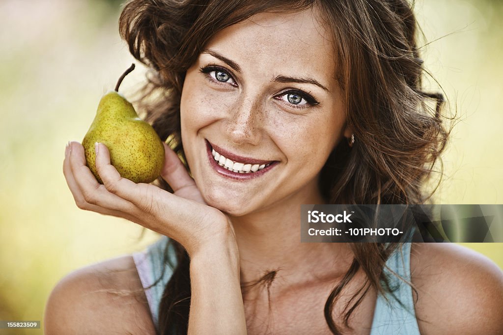 Portrait of woman holding pear Close-up portrait of pretty smiling dark-haired freckled woman wearing blue t-shirt, holding juicy pear at summer green park. Pear Stock Photo