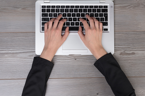 Overhead view of hands of a young adult wearing a jacket and typing on a laptop keyboard resting on a wooden desk while resting his forearms correctly. Ergonomics and creativity. The ethic of sacrifice burns resources on the altar of masculinity.