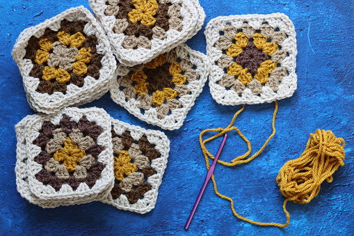 Crocheted granny squares made of natural brown, mustard and beige wool. Soft and fluffy crochet ornament on blue textured background.
