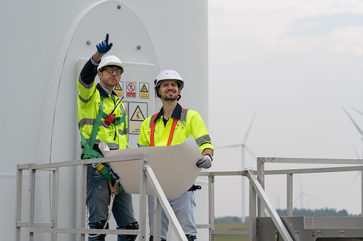 Two engineer in uniform with helmet safety pointing and holding blueprints to inspection and maintenance of wind turbine in wind farms to generate electrical energy, Renewable power energy.