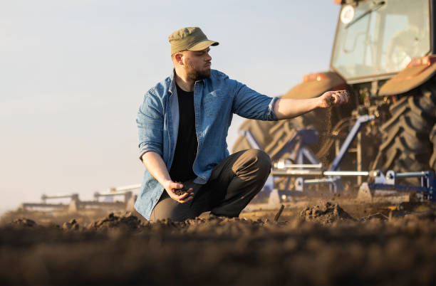 istock Farmer examing dirt while tractor is plowing field 1558296615