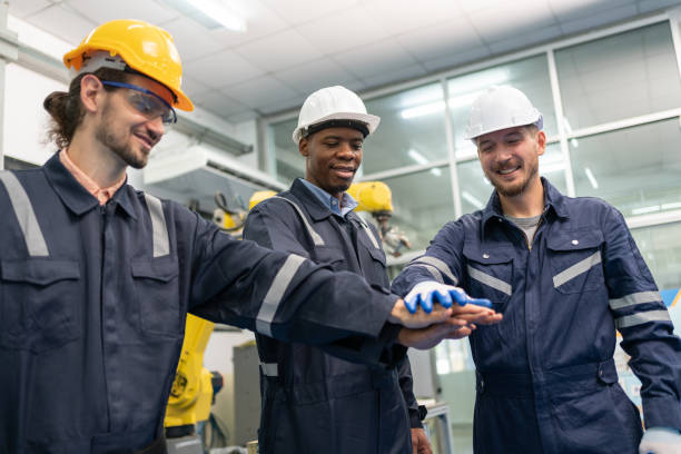 gruppe von roboteringenieuren trägt helme im stehen und reicht sich an den händen, um in der fabrikwerkstatt einsatzbereit zu sein. techniker, professionelles team, roboterarmschweißmaschine. - welder manual worker african descent steel worker stock-fotos und bilder