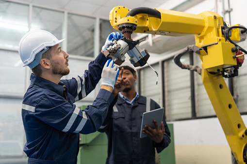 Male engineers wearing vest with helmet safety repairing robot arm welding machine in factory. Team of technician using tool and laptop working at robot industrial.
