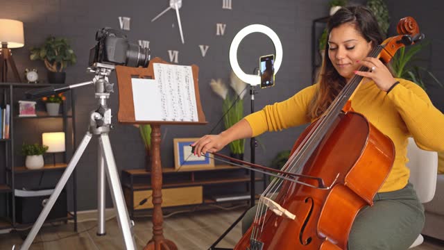 Young woman filming herself while playing violoncello at home