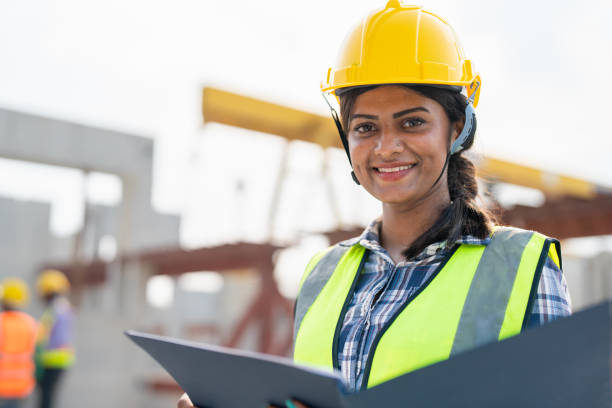 istock Asian woman engineer holding document smiling at construction site. Confident female Indian wearing protective helmet and vest working in factory marking precast concrete wall for real estate housing. 1558333992
