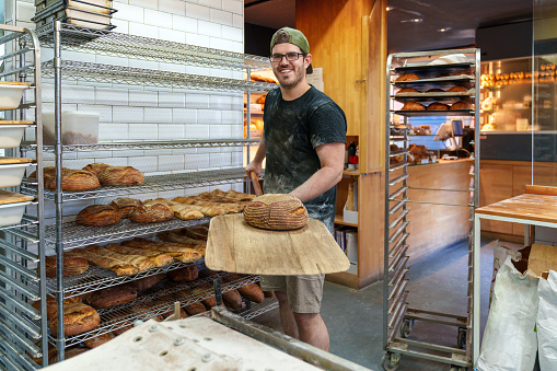 A joyful baker in a bustling bakery, armed with a bread peel, stands by a cart filled with freshly baked loaves, ready for the oven.