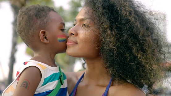 Little black 4 years old boy who painted his face with LGBTQ flags, kissing his mother's cheek at city street during marriage equality pride celebration. LGBTQ African American mother with afro hair style hugging her child boy during enjoying street carnival LGBTQ parade.