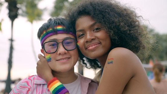 LGBTQ lesbian couple, Asian short hair and African American female with afro hairstyle with pride flag on their faces for love, equality and support during joining with the pride event in city center. Gay, lesbian couple hugging each other to support marriage equality for human rights parade event.
