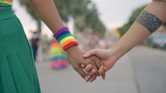 Close up of couple lesbians' hand holding each other's hands while joining the pride event in city center. A happy gay Asian with tattoo and African American with rainbow wristbands couple are street outdoors walking to support marriage equality for human rights among parade street celebration.