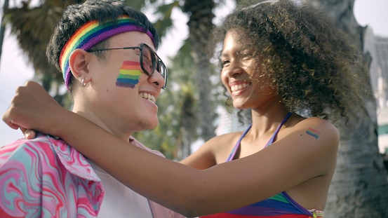 Two mid adult Hispanic women at a pride event, embracing and looking each other. They are participating in a LGBTQI Pride event. They are smiling. LGBTQ lesbian couple hugging each other to support marriage equality for human rights among parade street celebration.