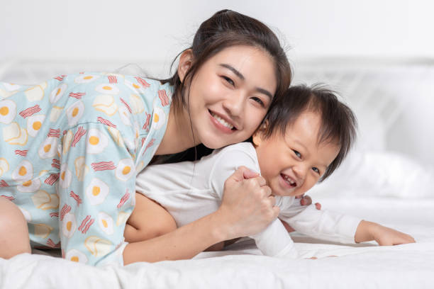 cute asian mom hugging her baby boy on bed smile and happiness together. little baby toddler sit on bed and relax with mother speding time to learning and growth. good momen of family time. mother day - love growth time of day cheerful imagens e fotografias de stock