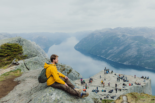 Side View of a male in yellow jacket with backpack sitting at the edge of the cliff looking at crowds of tourists on the cliff with view of beautiful fjord with mountains in South Norway, Scandinavia