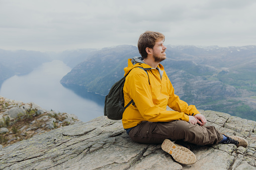 Side View of smiling male in yellow jacket with backpack sitting at the edge of the clip looking at beautiful fjord with Mountain View in South Norway, Scandinavia
