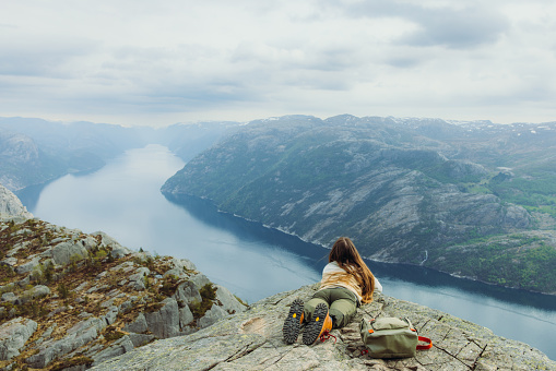 Rear View of happy female hiker lying down on scenic Preikestolen mountain looking at scenic view of beautiful fjord from above