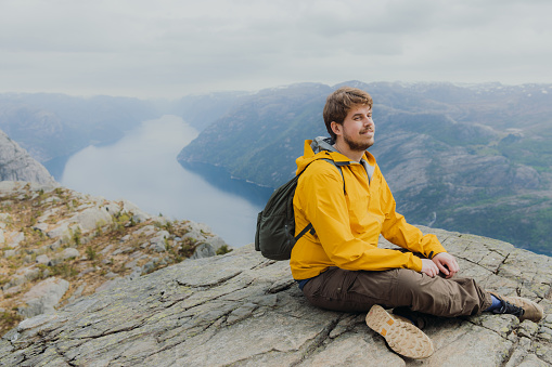 Side View of smiling male in yellow jacket with backpack sitting at the edge of the clip looking at beautiful fjord with Mountain View in South Norway, Scandinavia