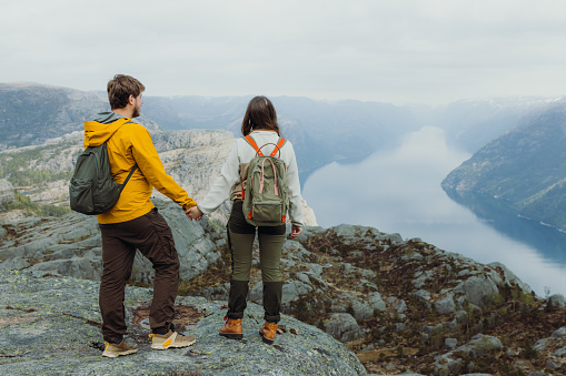 Rear View of heterosexual couple of backpackers getting to the top of the mountain admiring a view of idyllic Lysefjord in South Norway, Scandinavia