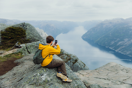 Side View of a male in yellow jacket with backpack using Internet while sitting at the edge of the cliff with idyllic view of Lysefjord in South Norway, Scandinavia