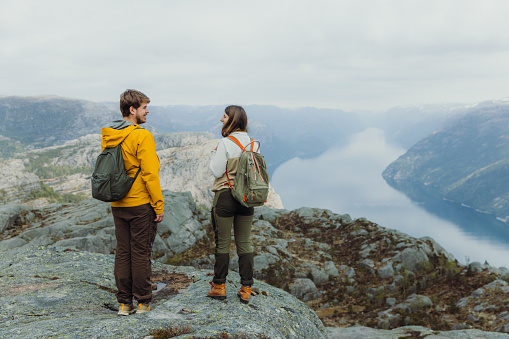 Rear View of happy heterosexual couple of backpackers getting to the top of the mountain admiring a view of idyllic Lysefjord in South Norway, Scandinavia