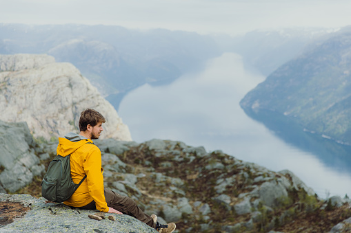 Side View of smiling male in yellow jacket with backpack sitting at the edge of the clip looking at beautiful fjord with Mountain View in South Norway, Scandinavia