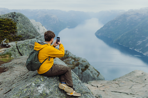 Side View of a male in yellow jacket with backpack using Internet while sitting at the edge of the cliff with idyllic view of Lysefjord in South Norway, Scandinavia