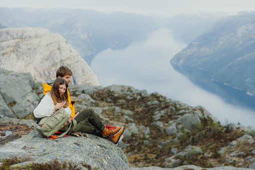 Side view of happy heterosexual couple relaxing on the cliff having lunch and looking at scenic view of mountain fjord in South Norway, Scandinavia