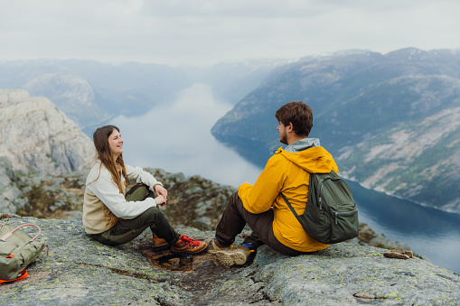Side view of smiling heterosexual couple relaxing on the top of the mountain and talking with background view of scenic mountain fjord in South Norway, Scandinavia