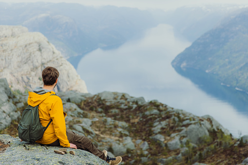 Side View of smiling male in yellow jacket with backpack sitting at the edge of the clip looking at beautiful fjord with Mountain View in South Norway, Scandinavia