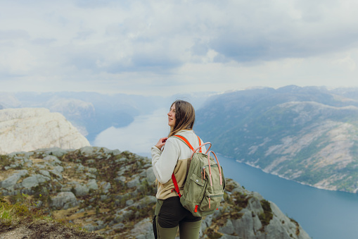 Rear View of smiling female with long hair and backpack hiking to the top of Preikestolen contemplating a view of scenic mountain fjord in South Norway, Scandinavia