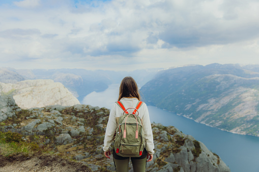 Rear View of smiling female with long hair and backpack hiking to the top of Preikestolen contemplating a view of scenic mountain fjord in South Norway, Scandinavia