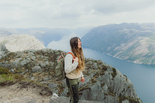 Portrait of smiling female with long hair and backpack hiking to the top of Preikestolen contemplating a view of scenic mountain fjord in South Norway, Scandinavia