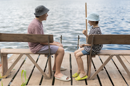 Two teenage boys in hats sitting on benche on the pier near lake.