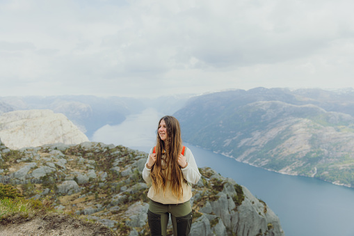 Portrait of smiling female with long hair and backpack hiking to the top of Preikestolen contemplating a view of scenic mountain fjord in South Norway, Scandinavia