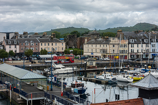 Rothesay, Scotland - 15th Juily 2023: View over part of the Marina and Esplanade of the seaside town of Rothesay on the Isle of Bute, Scotland. Pleasure craft can be seen in the marina, the Cabmen's  Rest (1874) is on the pier, a Hop-on hop-off sightseeing bus is at the bus stop, the shops and businesses of the promenade can be seen and Rothesay Castle (ruins) is in the background.