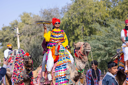 Bikaner, Rajasthan, India - January 13 2023: Camel Festival, Portrait of an young rajasthani male with beard and moustache wearing white traditional rajasthani dress and turban riding on camel cart.
