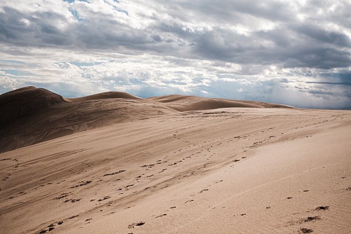A gorgeous view of sand dunes in a desert under a cloudy sky, dotted with footprints